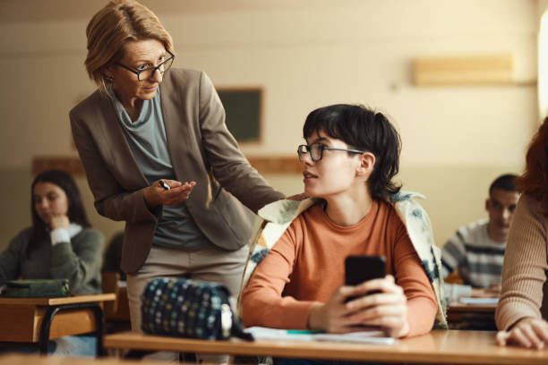 Displeased senior teacher talking to her student who is using a mobile phone during a class in the classroom.